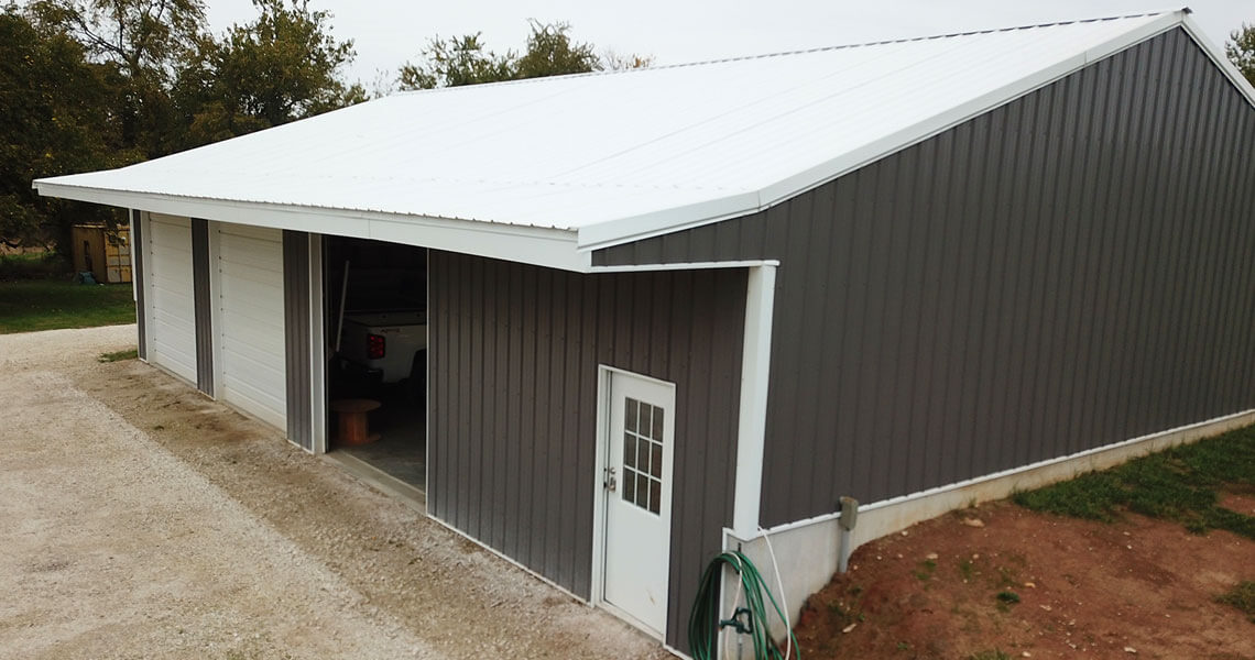 A large metal garage with gray walls and a white roof featuring three garage doors and a single entry door on a gravel driveway, surrounded by green grass and trees in the background.
