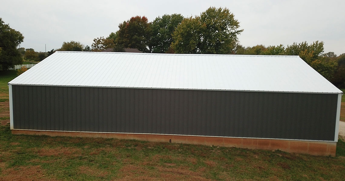 A large, rectangular metal building with a gray exterior and a white roof stands on grassy land. Trees with green and autumn-colored leaves are visible in the background under an overcast sky.