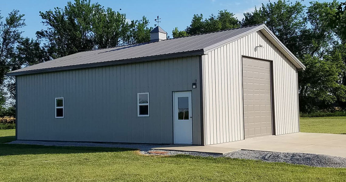 A small grey metal building with a sloped roof stands on a concrete pad. It has two small windows, a white door, and a large beige garage door. Trees are visible in the background under a clear blue sky.