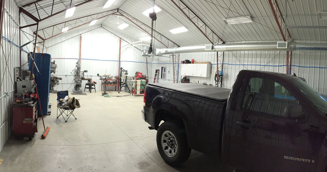 A large workshop with metal walls and a high ceiling features various tools and machinery along the sides. A black pickup truck is parked inside, and a foldable chair is nearby. The space is well-lit with overhead lights.