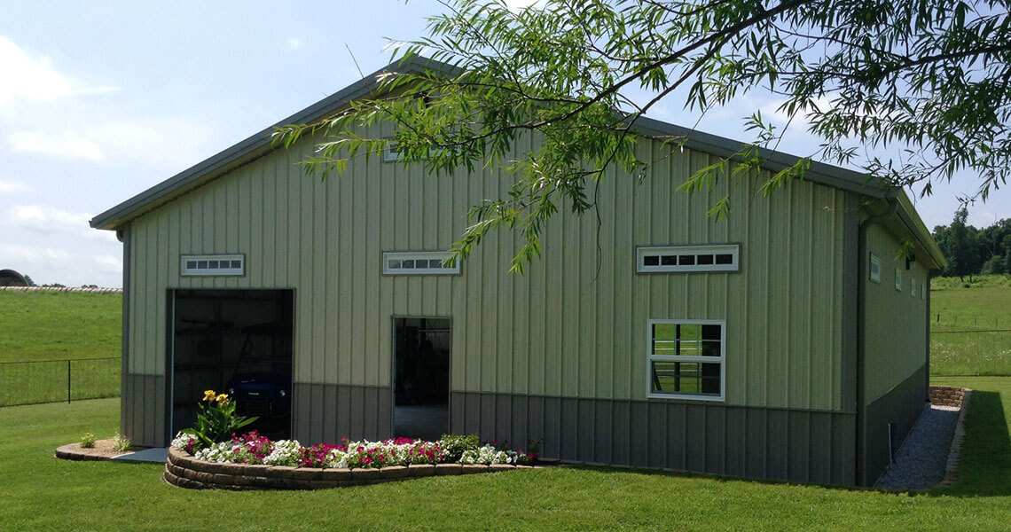 A large green metal barn with small rectangular windows and two open garage doors, surrounded by a lush field. There's a flower bed with colorful blooms in front of the barn and a leafy branch overhead.