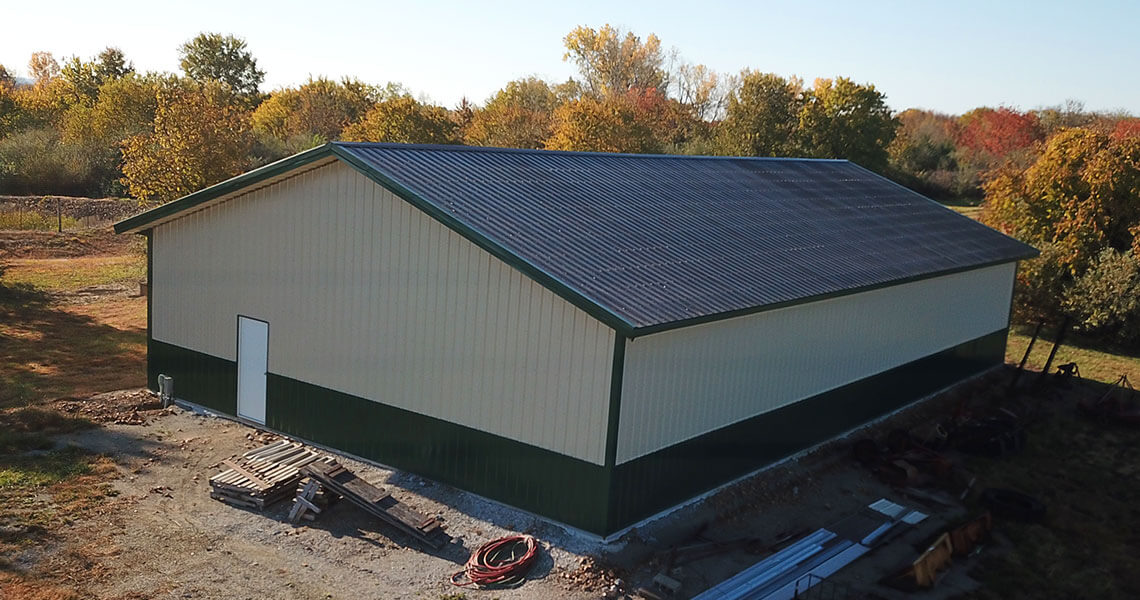 A large, metal-roofed storage building with beige walls and green trim is situated in a wooded area. The surrounding trees are filled with fall foliage. Various construction materials and equipment are scattered nearby.