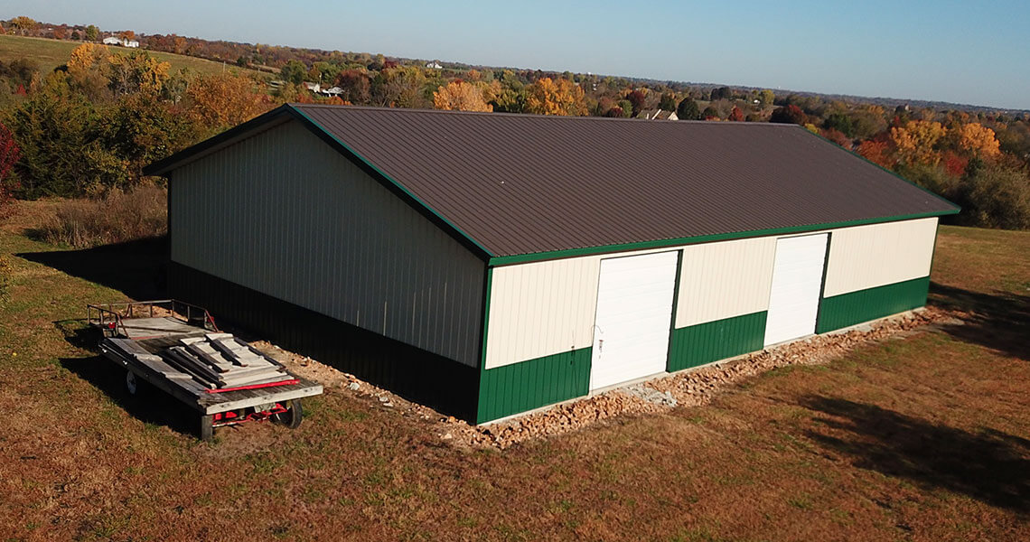 A large, rectangular barn with a dark roof and green and cream siding stands in a grassy area. Two closed white garage doors are on the side. Nearby is a flatbed trailer with stacked lumber. In the background, colorful autumn trees are visible.