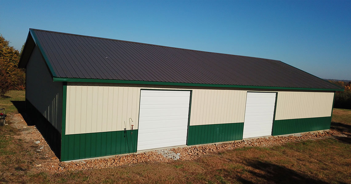 A large pole barn with cream-colored siding, green trim, and a dark roof stands in an open grassy area under a clear blue sky. It has multiple white garage doors on one side.