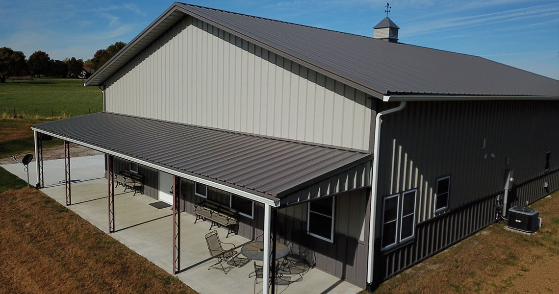 A large, modern barn with a sloped roof and covered porch is set in a grassy area. It features metal siding and windows on the side. Patio furniture is visible on the concrete porch, and trees are seen in the distance under a blue sky.