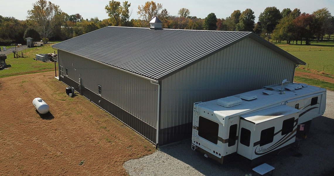A large metal building with a gray roof stands beside a parked RV. There is a propane tank on the grass nearby, and trees with fall foliage in the background under a clear sky.