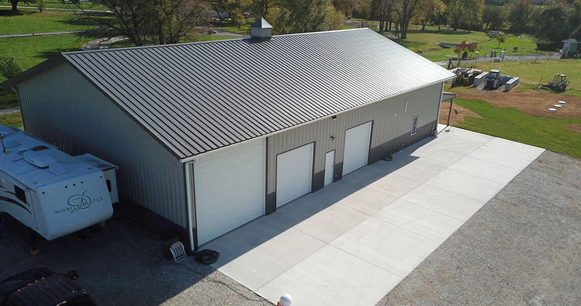 Aerial view of a large metal building with a sloped, dark grey roof and three garage doors, surrounded by a concrete driveway. A parked RV trailer is visible on the left side, and there are green fields and trees in the background.