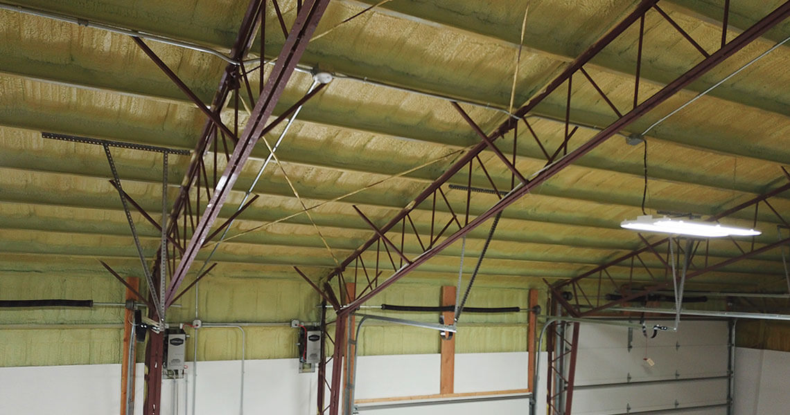 View of an insulated industrial warehouse ceiling with visible steel beams and supporting structures reminiscent of metal garage kits. Fluorescent lights hang from the ceiling, illuminating a partially visible garage door below.