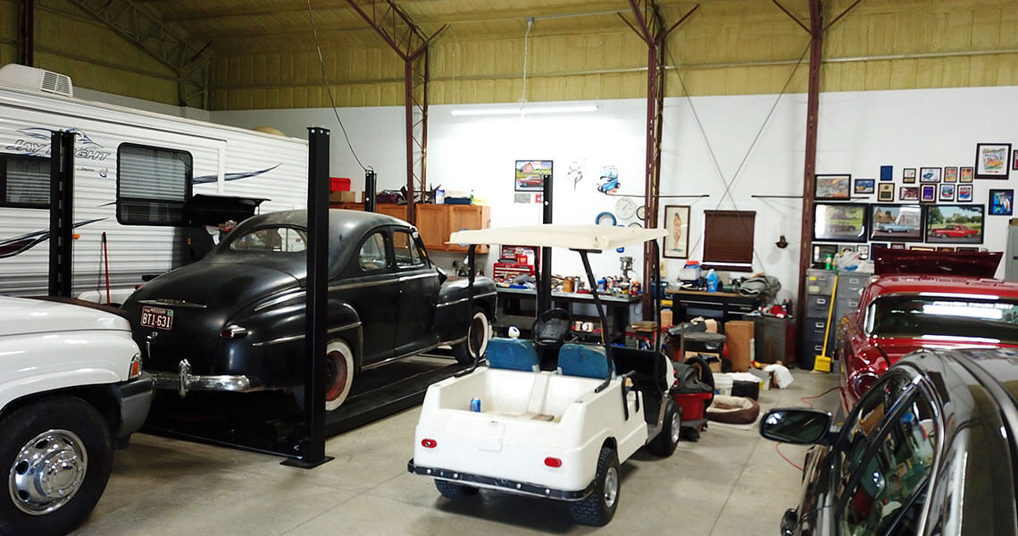 A cluttered garage with a classic black car on a lift, a white golf cart, and various tools and equipment. A camper is parked on the left. Posters and framed pictures adorn the walls. The space has a workshop feel.