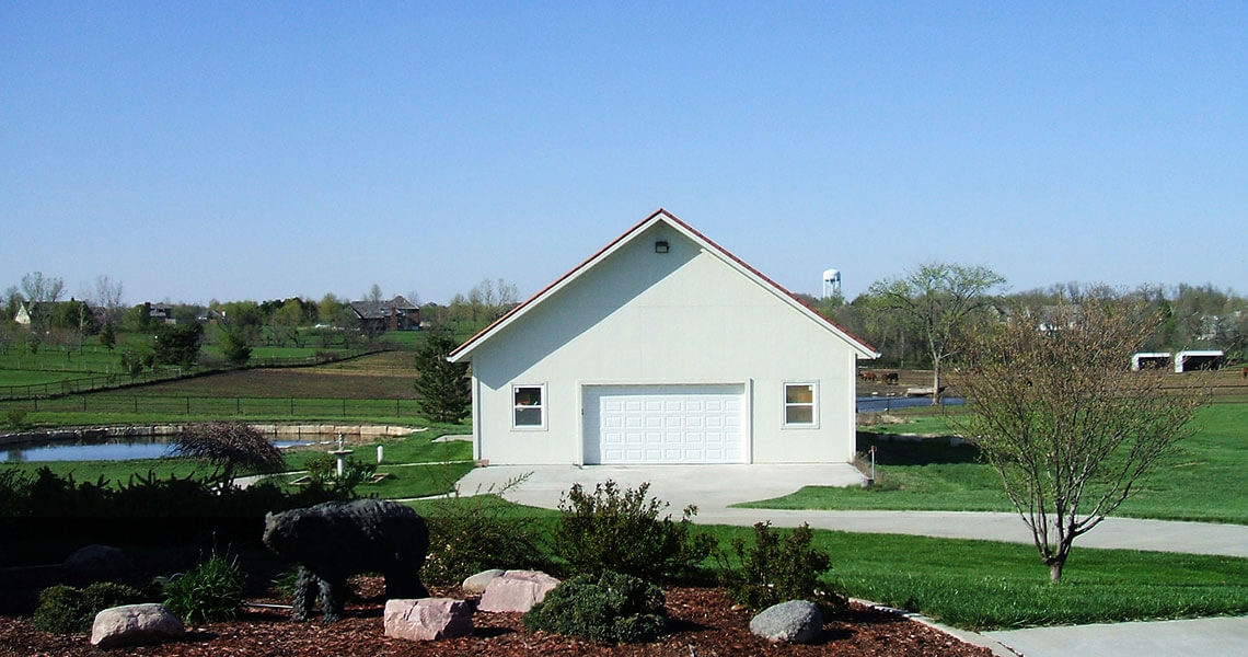 A white barn with a large garage door sits on a grassy landscape. In the foreground, there are shrubs and rocks, and a pond is visible to the left. Trees and sparse buildings are in the background under a clear blue sky.
