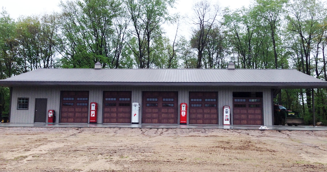 A rectangular building with a metal roof evokes nostalgia as it houses four vintage red and white gas pumps reminiscent of an old Texaco Station, set in front of four wooden garage doors. Trees form a backdrop, and the ground is a mix of dirt and gravel, completing this Laviola scene.