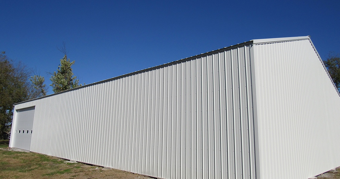 A large white metal warehouse building with a sloped roof stands under a clear blue sky, reminiscent of a modern barn. Hess Farms surrounds the structure with grass and sparse trees, crafting a serene rural or industrial setting.