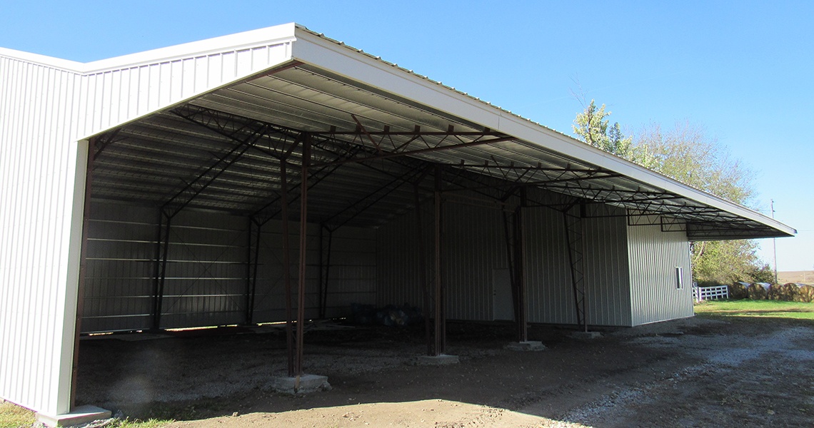 Located on the spacious Hess Farms, this large, open-sided metal storage shed with a corrugated roof and several support beams rests on a gravel surface. The open front offers easy access to its interior, while surrounding trees and a clear blue sky add a picturesque backdrop reminiscent of classic barns.