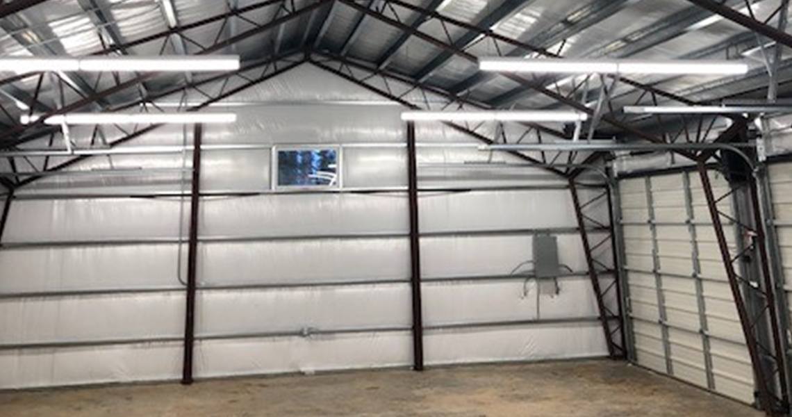The Centennial Garage boasts an interior of an empty metal warehouse, featuring white insulated walls and rafters. Fluorescent lights illuminate the space, perfect for automotive repair. A closed garage door stands beside a small window near the roof peak, overlooking the concrete floor.