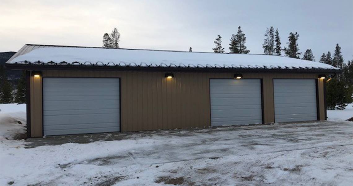 Centennial Garage stands with its three closed, light gray doors under a sloped metal roof. Snow blankets the roof and ground, while evergreen trees peek through the overcast sky. Warm exterior lights glow above each door, casting a welcoming ambience amid the winter chill.