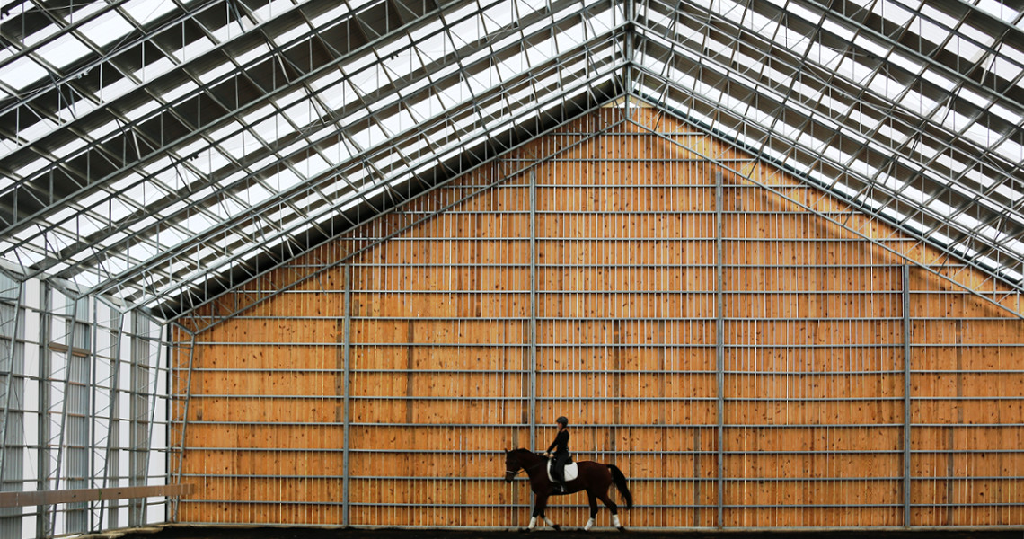 A person on horseback inside a large, wooden-structured riding arena with a high metal roof and grid-like pattern. The setting in Maine is spacious, with the rider and horse perfectly centered against the backdrop of the wall.