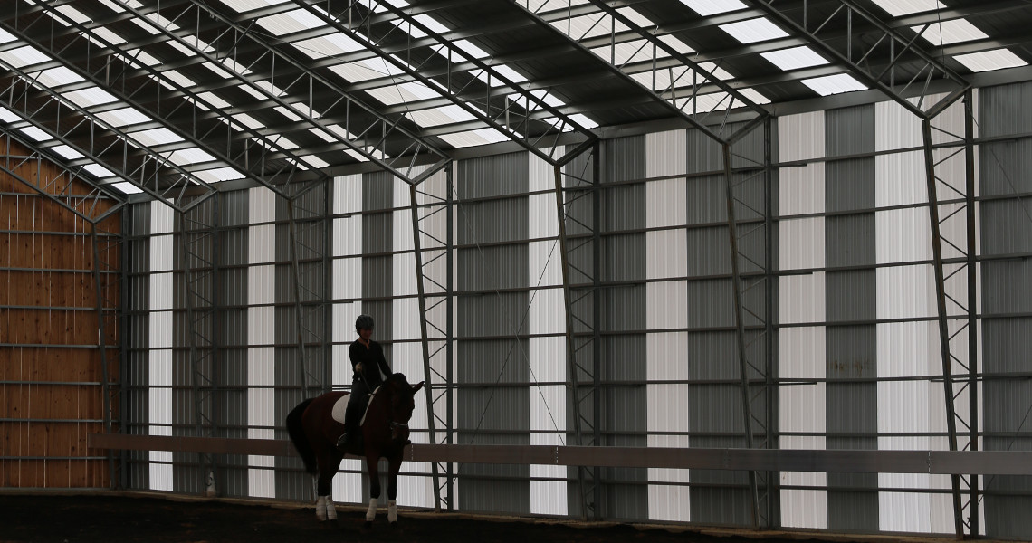 A person rides a horse inside a spacious riding arena in Maine, featuring a high, metal-framed ceiling and walls. The space is bright, with natural light streaming through translucent panels in the roof.