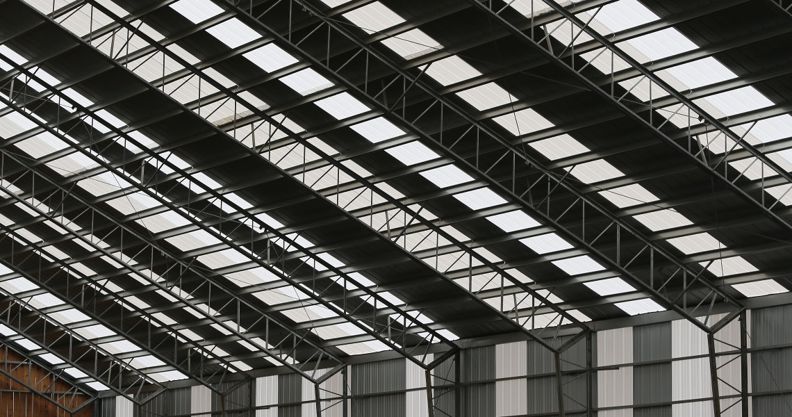 The interior view of a large industrial warehouse resembles an arena with its high ceiling featuring metal beams and skylights. Vertical panels in varying shades of grey adorn the walls, while natural light elegantly filters through the roof, evoking the expansive skies of Maine.