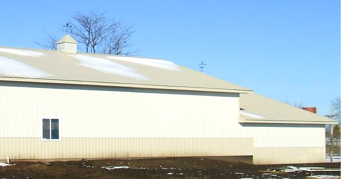 A large beige barn with a metal roof stands against a blue Maine sky, resembling a rustic arena. Snow patches are visible on the roof and ground, while a leafless tree and distant silo complete the serene scene.