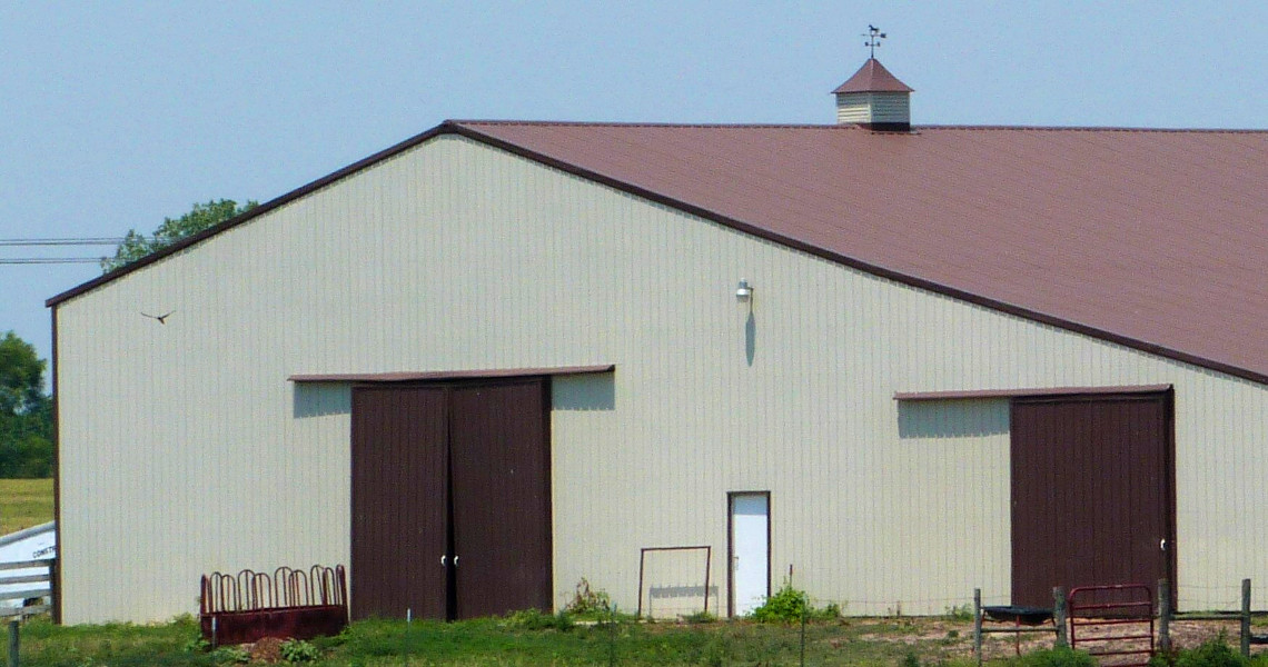 A large beige barn with a red roof graces the landscape of Riverview Farms, standing proudly against a clear blue sky. Its two large brown sliding doors are complemented by a small metal rooster weather vane atop the roof, while farming equipment sits prominently in the foreground.
