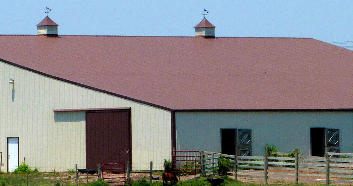 At Riverview Farms, a large barn with a brown roof and two cupolas sits in a rural setting. The building features two grand doors and is encircled by a wooden fence. Scattered nearby are some small items and a horse, all under a clear blue sky.