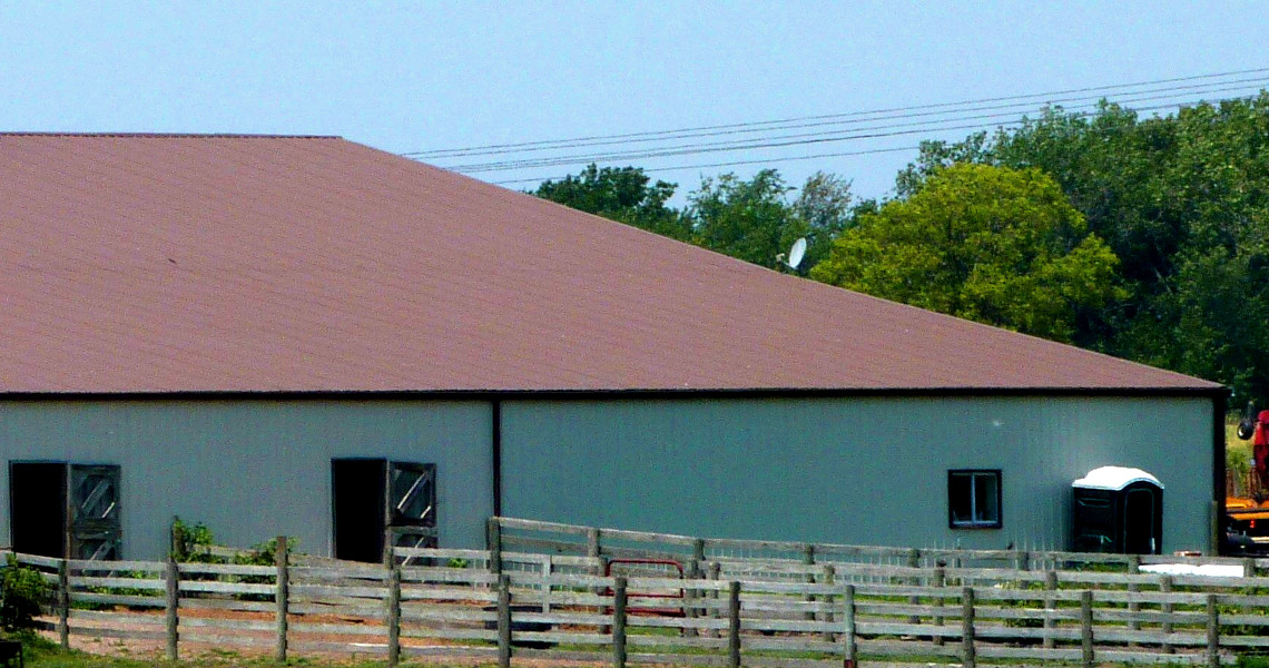 Riverview Farms features a large barn with a reddish-brown roof standing amidst a rural setting. Fenced paddocks grace the foreground, while trees frame the idyllic scene in the background.