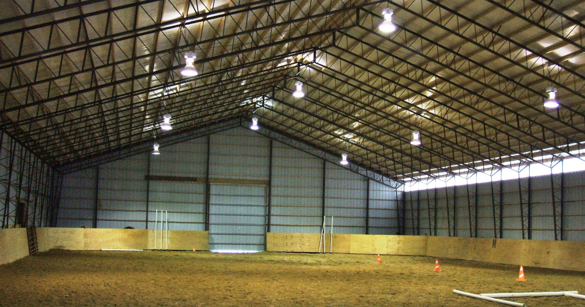 The image captures the interior of Riverview Farms' spacious indoor riding arena, featuring high metal beams and a dirt floor. The space is beautifully illuminated by overhead lights, with barriers and several orange cones strategically placed near the walls.
