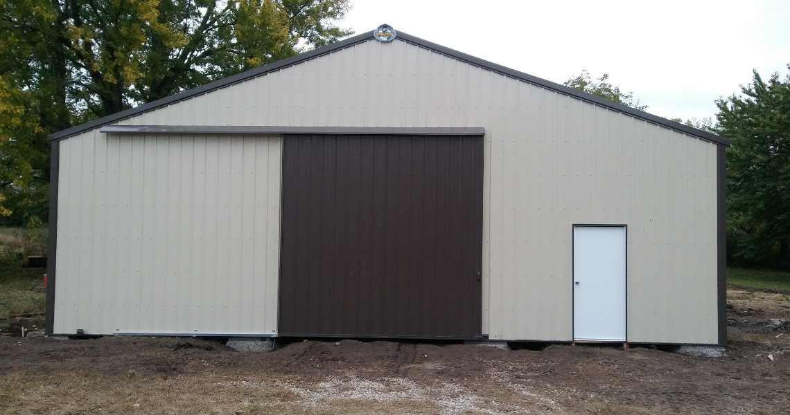 A beige and brown metal storage building with a large sliding door on the left and a regular door on the right. The structure is set in a clearing with trees visible in the background. The ground is bare soil in front of the building.