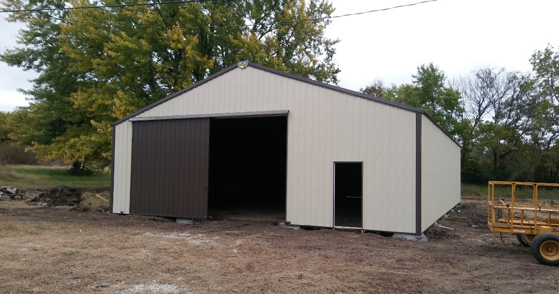A large, beige metal barn with a dark brown roof and sliding doors stands on a patch of dirt and grass. The barn door is partially open, revealing the interior. Surrounding trees display autumn foliage. A yellow cart is parked nearby.