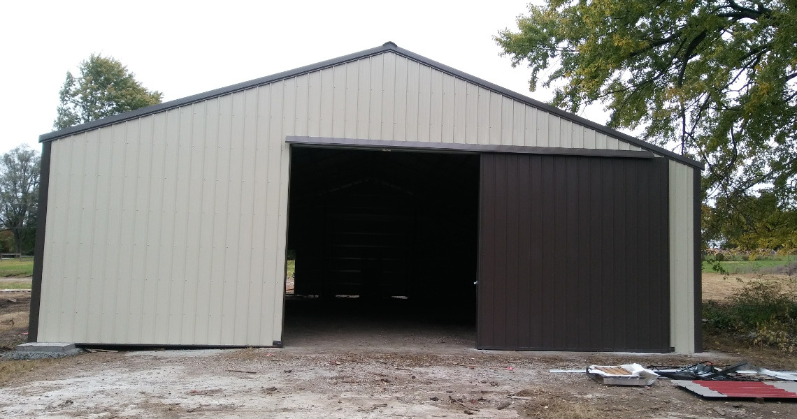 A large metal barn with one open sliding door, showing a dark interior. The structure is surrounded by a grassy area, with trees visible in the background. Building materials are scattered on the ground nearby.