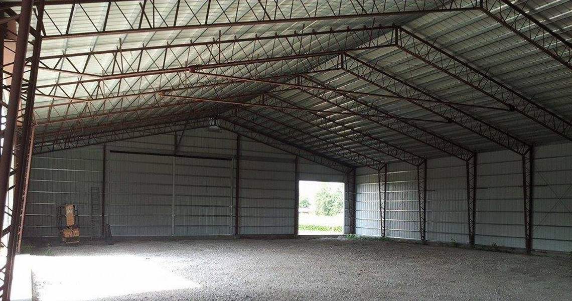 Interior of an empty metal warehouse with a high ceiling and open sliding door. The structure features exposed steel beams and a gravel floor, with sunlight streaming through the entrance, illuminating the interior space.