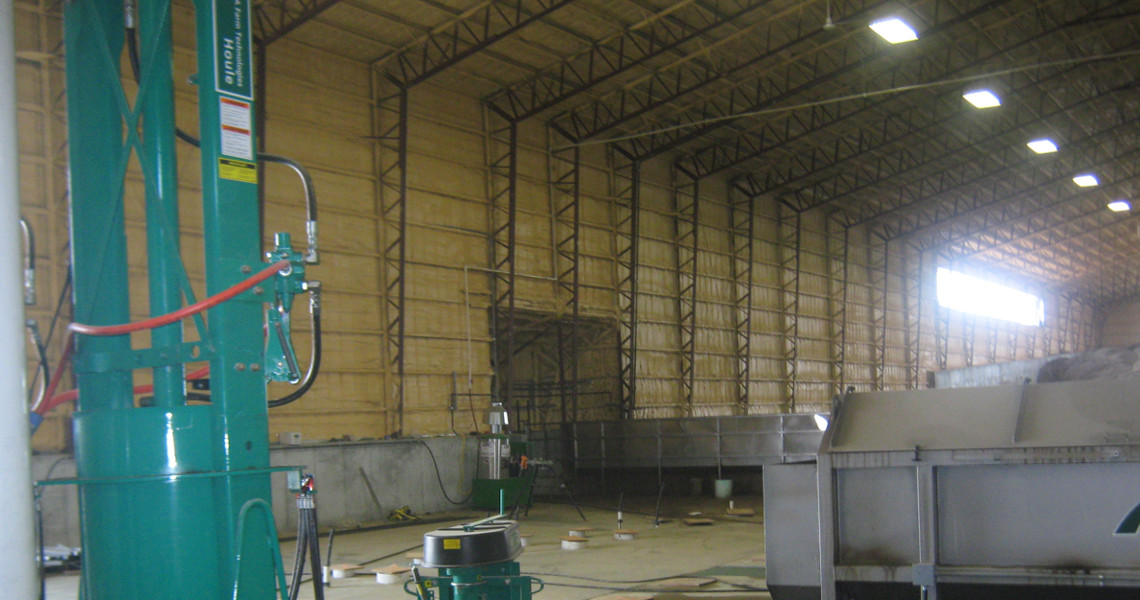 Interior of a large industrial warehouse with high ceilings and metal beams. The space contains green machinery and a few scattered items on the floor. Bright overhead lights and a large window illuminate the area.