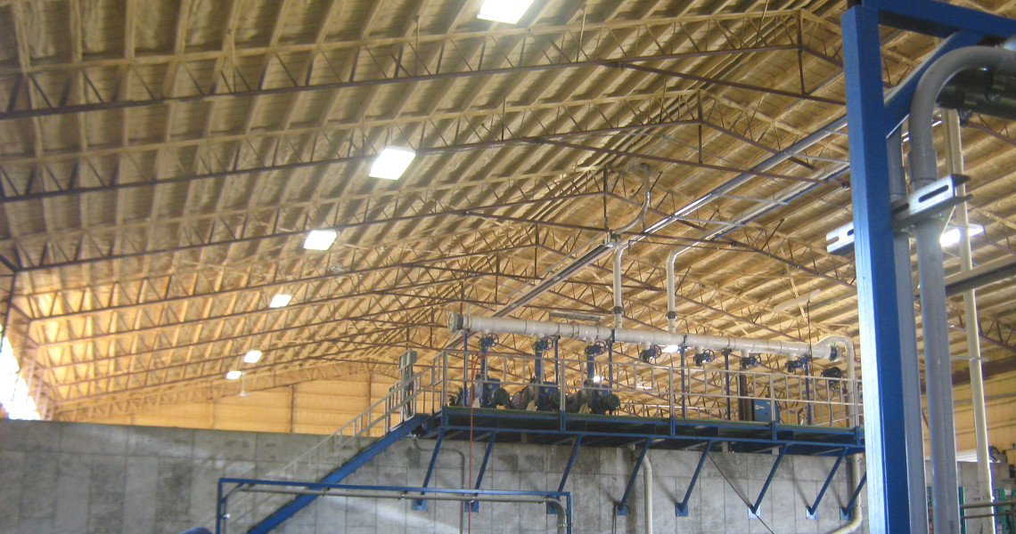 Interior view of an industrial facility with a high ceiling and steel beams. Bright lights illuminate the structure. A blue metal framework and a concrete wall are visible, with pipes and ducts running through the space.