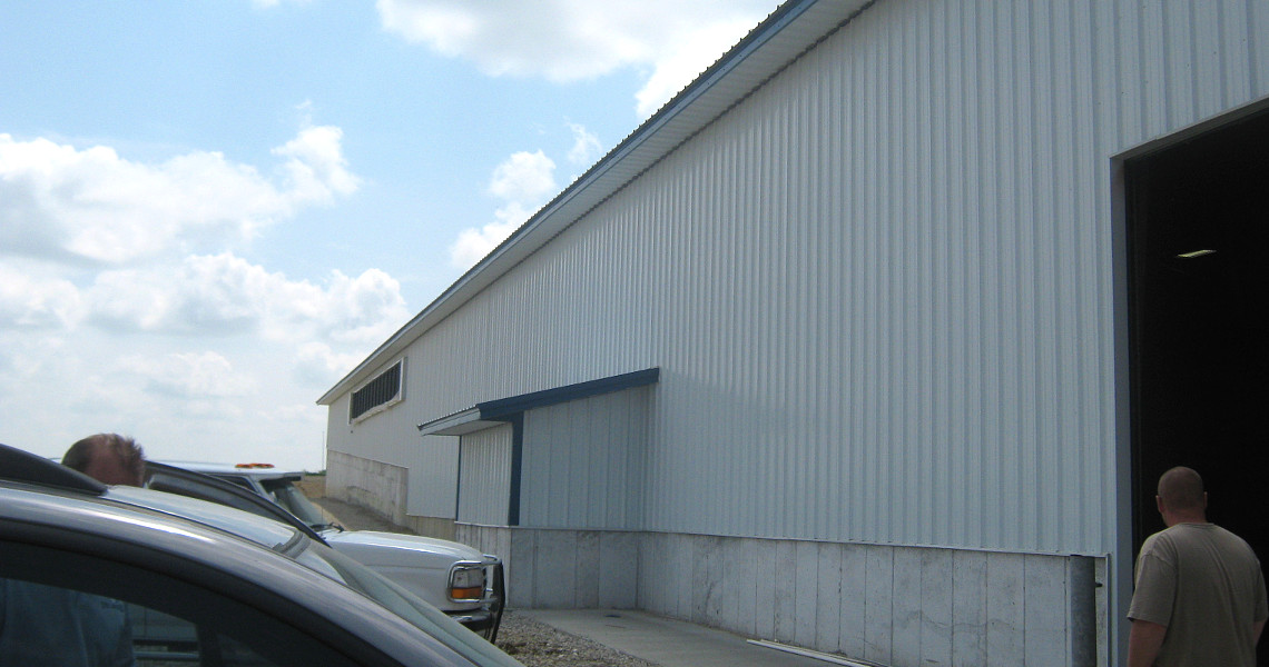 A large white industrial building with a sloped roof stands against a cloudy sky. Two vehicles are parked in the foreground, and a person is walking toward a doorway on the right.