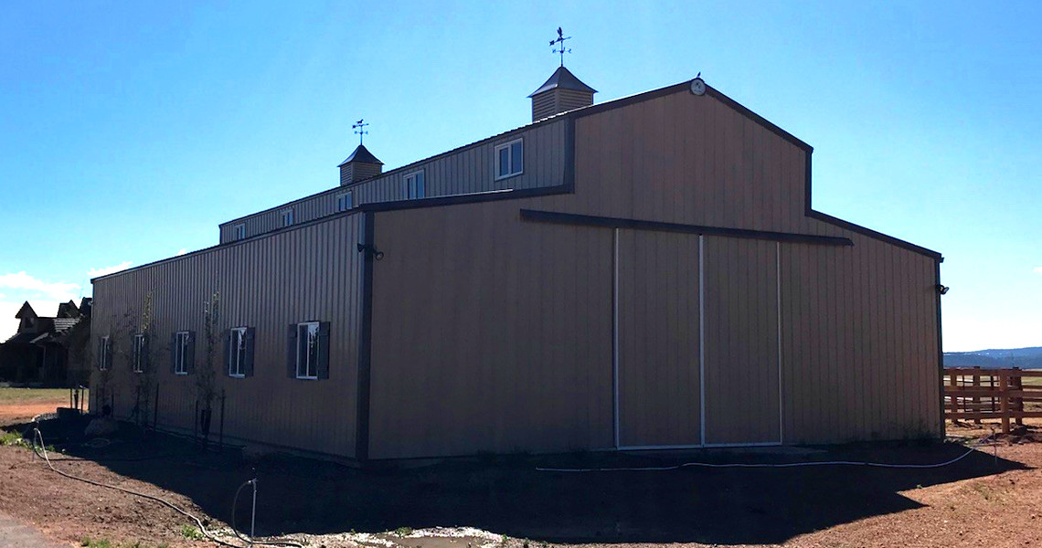A large, tan metal barn with two cupolas and weather vanes on the roof stands under a clear blue sky. The structure features several small windows and a large sliding door. The surrounding landscape is dry and sparsely vegetated.