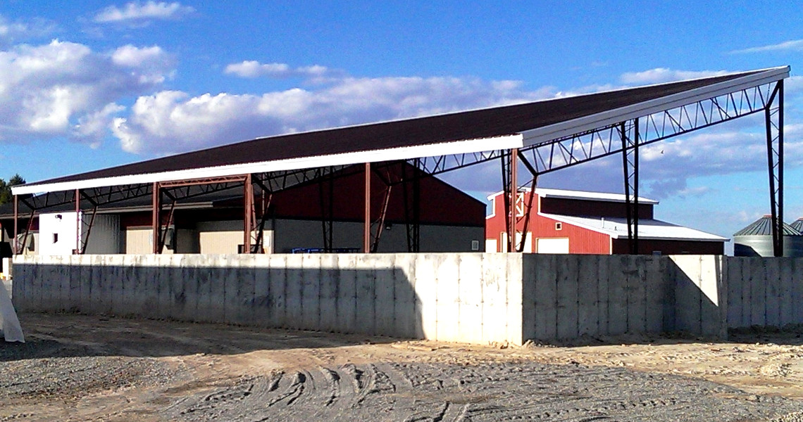 A large agricultural building with a sloped roof and open sides, surrounded by a concrete wall, under a blue sky with clouds.
