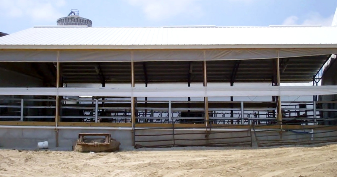 Exterior view of a large, open-sided barn or agricultural building with a white metal roof. The partially covered sides reveal metal framework and fencing inside. A silo is visible in the background, with bare ground in the foreground.