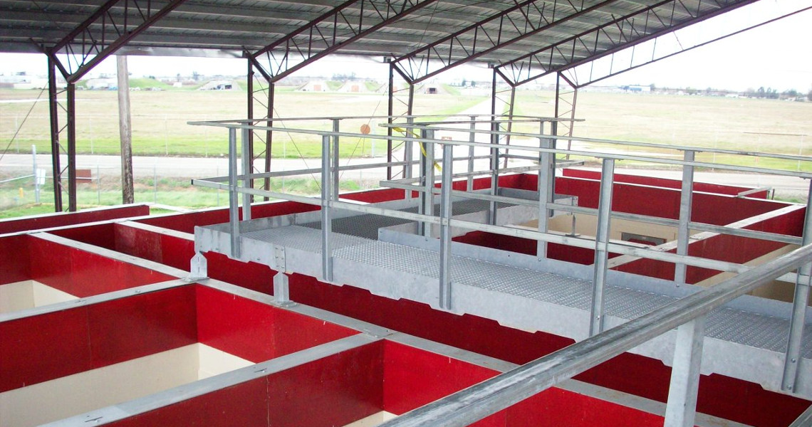 View of a large industrial building's interior with an open framework. Red and white sections are visible below a metal walkway and railing, under a large roof with supporting beams. The surrounding landscape is visible through the open sides.
