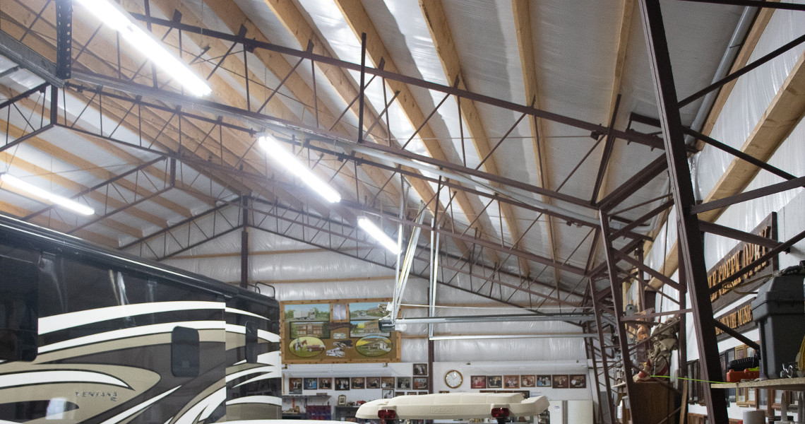 Interior of a spacious garage or workshop with a high ceiling and metal beams. A large RV, ideal for a Kansas getaway, and a covered vehicle are parked inside. Various items and framed pictures adorn the walls, while fluorescent lights illuminate the space.