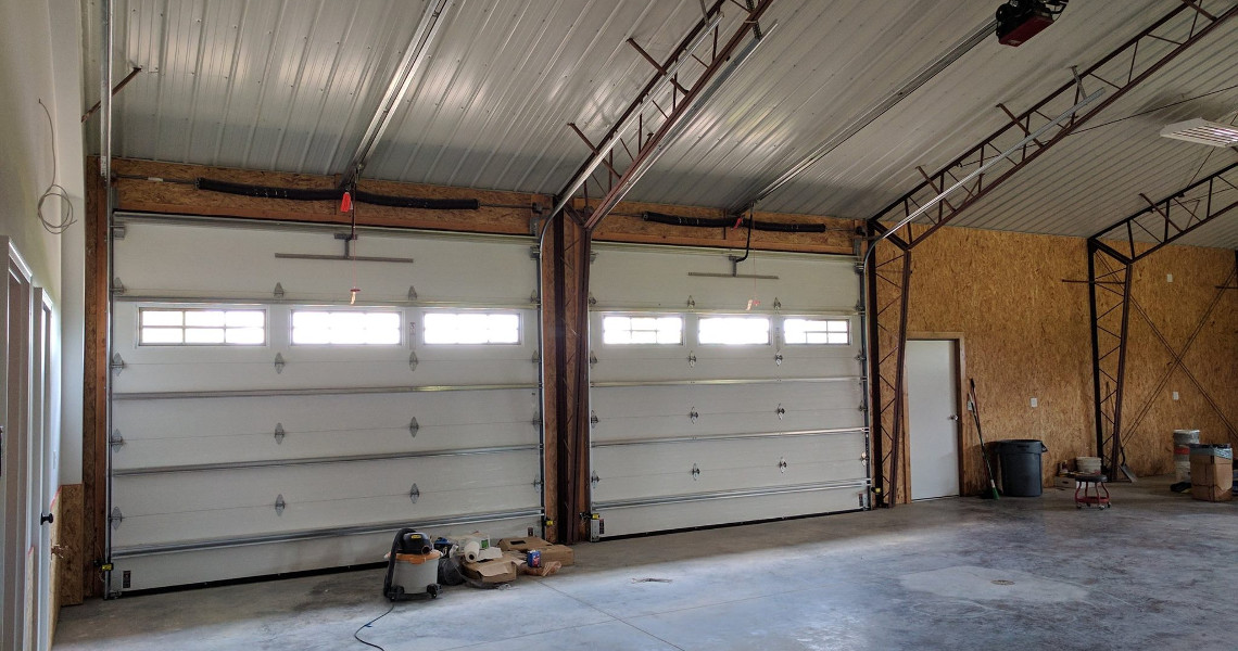 Inside the spacious garage of this Duncan Dream Home, two large, closed white garage doors with small windows stand out. The walls remain unfinished and the concrete floor is scattered with various tools and equipment, including a vacuum cleaner and buckets.