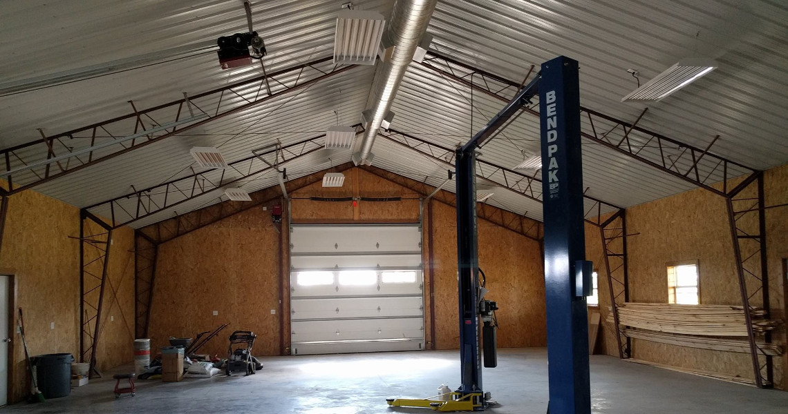 The interior of a large, empty garage or workshop in a Duncan dream home features a metal truss ceiling, plywood walls, and a BendPak car lift. This ideal space includes a garage door with various tools, equipment, and wood neatly piled on the sides.
