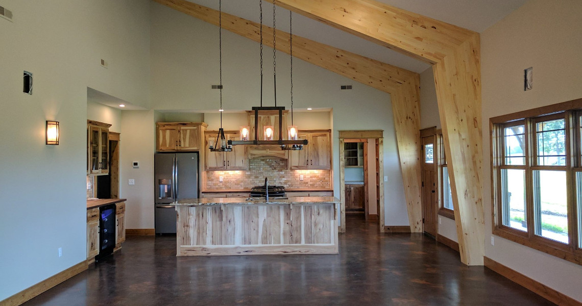 A dream kitchen with rustic wooden beams and cabinetry, featuring a modern island with pendant lights, stainless steel appliances, and a stone backsplash. Large windows and polished concrete floors give this Duncan-inspired space an open and airy feel.