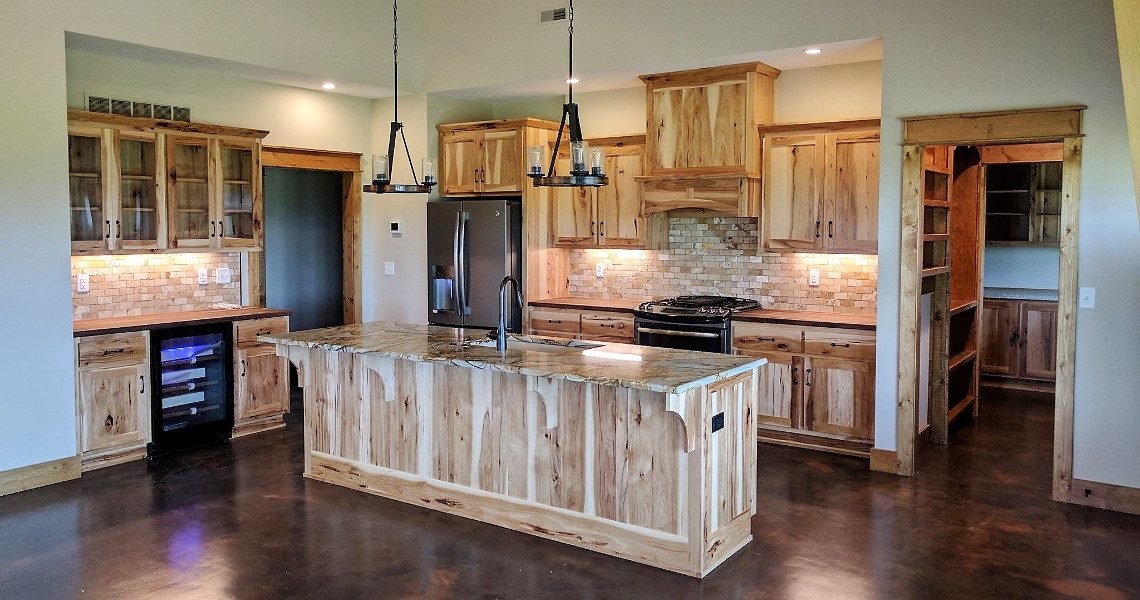 This dream kitchen features wooden cabinets, a large island with a marble countertop, and stainless steel appliances. Two pendant lights hang above the island, creating a welcoming home atmosphere. Shelving and a wine cooler are on the left, with a walk-in pantry to the right.