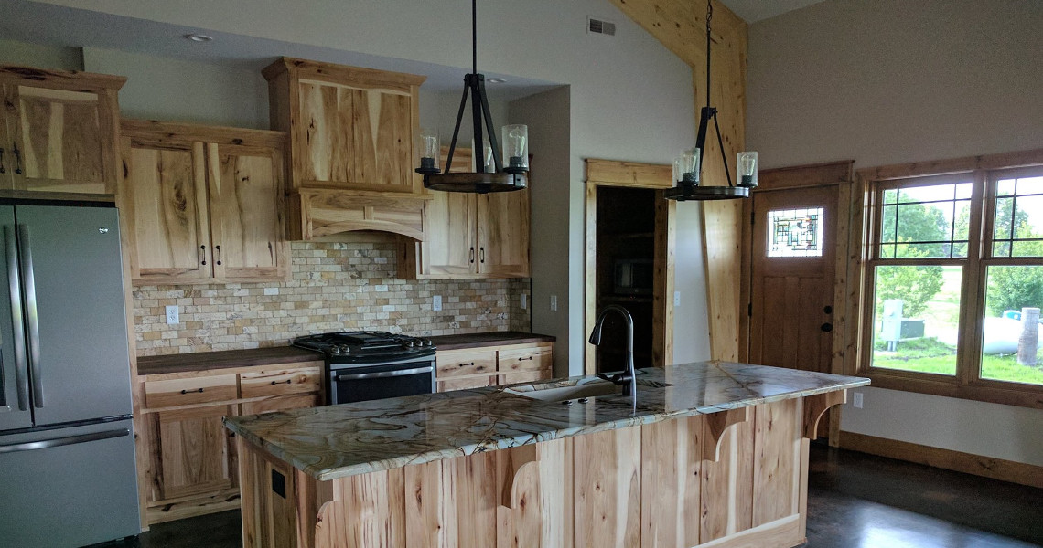 The Duncan Dream Home's rustic kitchen boasts wooden cabinets and a large island with a natural stone countertop. Two pendant lights illuminate the space, complemented by stainless steel appliances and a window framing lush greenery outside.