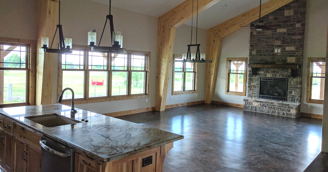 Welcome to your Duncan Dream Home: a spacious open-plan room featuring a stone fireplace, elegant wooden beams, and windows that flood the space with natural light. A kitchen island with a marble countertop and modern sink resides gracefully under sleek black pendant lights.