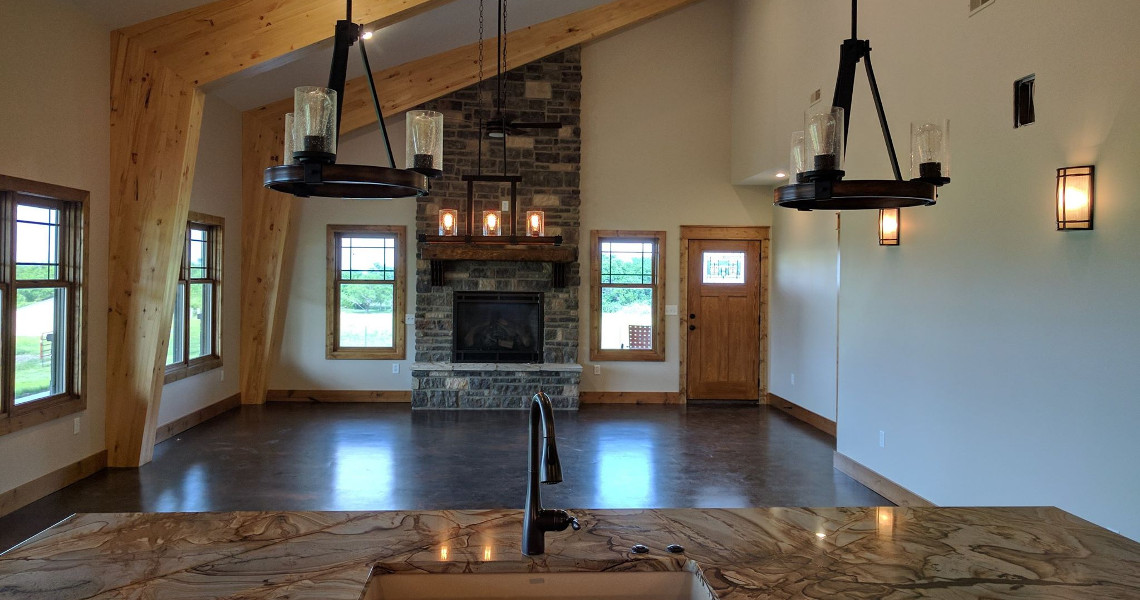 The image showcases a dream home with a rustic living room, marked by tall ceilings and exposed wooden beams. On one side, a stone fireplace complements large windows, while Duncan's modern chandeliers and a marble kitchen island grace the foreground.