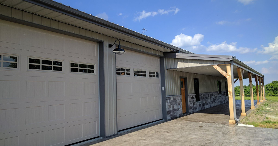 A modern barn-style dream home with large garage doors, featuring a wooden door and stone accents. The structure has a covered porch supported by sturdy wooden posts. The sky is clear and blue, with some scattered clouds—perfect for Duncan to call his own sanctuary.