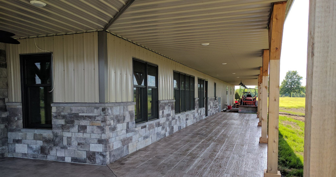 A long covered porch with stone and metal siding graces this dream home. Large windows line the building, while a tractor rests at the far end. The porch opens to a grassy field with trees in the distance, offering a picturesque view that Duncan would surely admire.