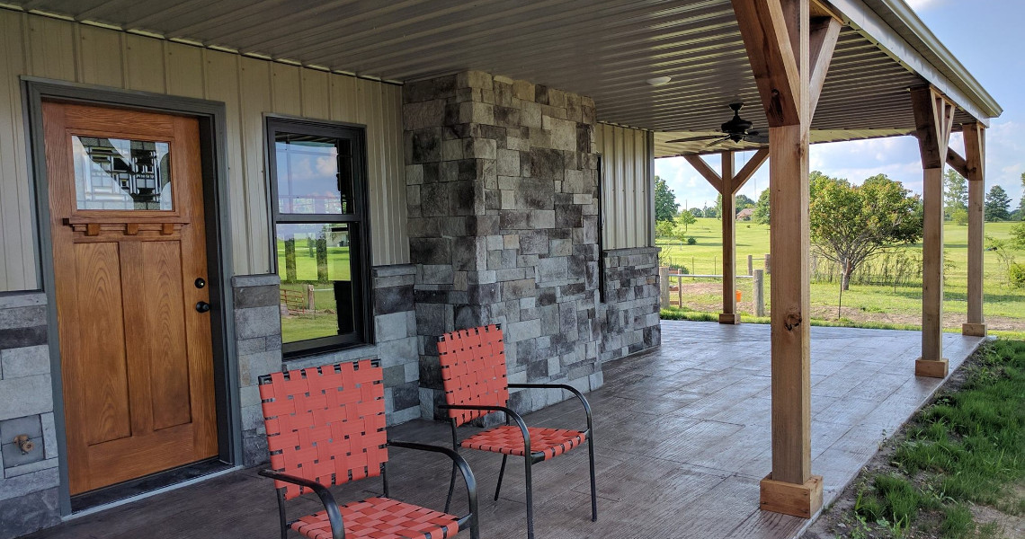 Step into your Duncan Dream Home: a covered patio featuring two orange woven chairs and stone wall accents alongside a rustic wooden door. Overlooking a grassy yard under partly cloudy skies, the area boasts sturdy wooden beams and a ceiling fan to complete this tranquil retreat.