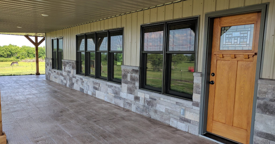 A covered porch showcases a wooden front door with decorative glass and large windows. The home's exterior blends beige panels and gray stone, offering a dreamy view of the green grassy field where a horse grazes in the distance.
